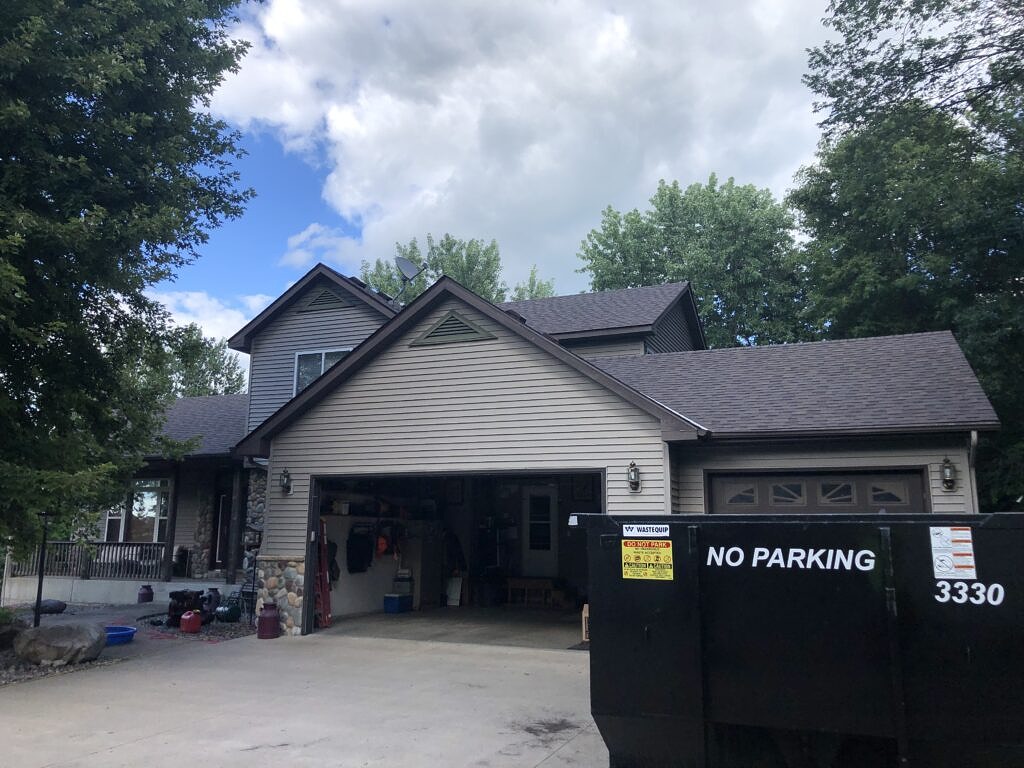 A tan house with new shingles with a roll off dumpster in the driveway.